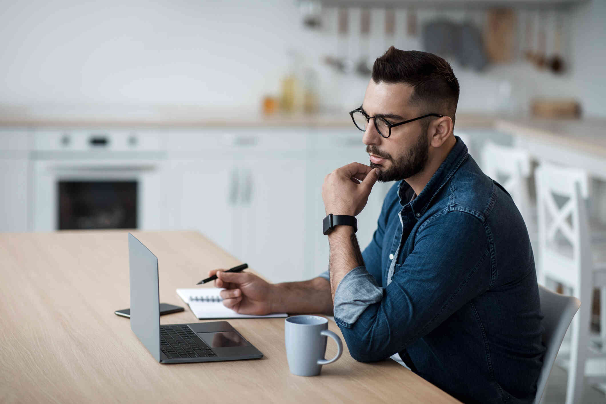 A man seated at a table appears focused on his laptop while taking notes.